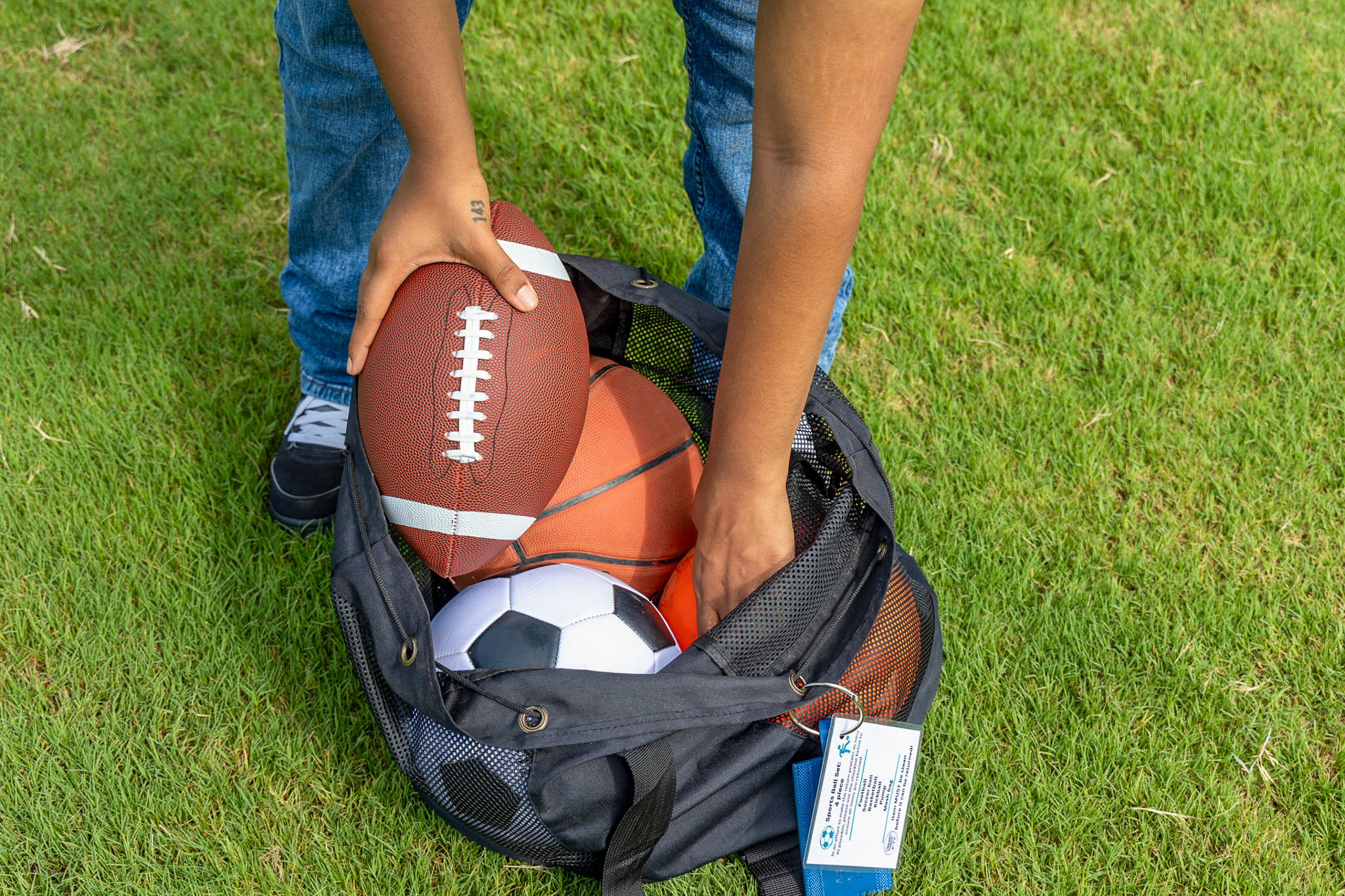 Person arranging athletic equipment in a black mesh bag on green grass.